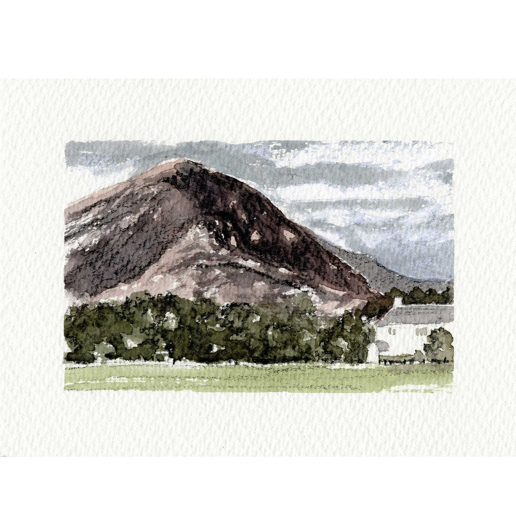 A watercolor landscape art print depicting a large dark mountain peak, green fields, trees, and a small white building under a cloudy sky, presented on a textured off-white paper background.